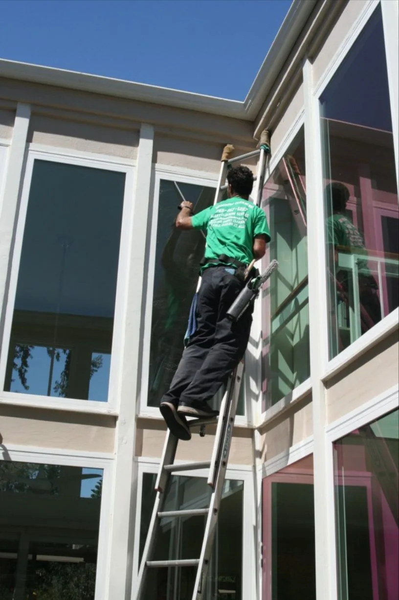 Technician in a green shirt on a ladder cleaning tall sunroom windows